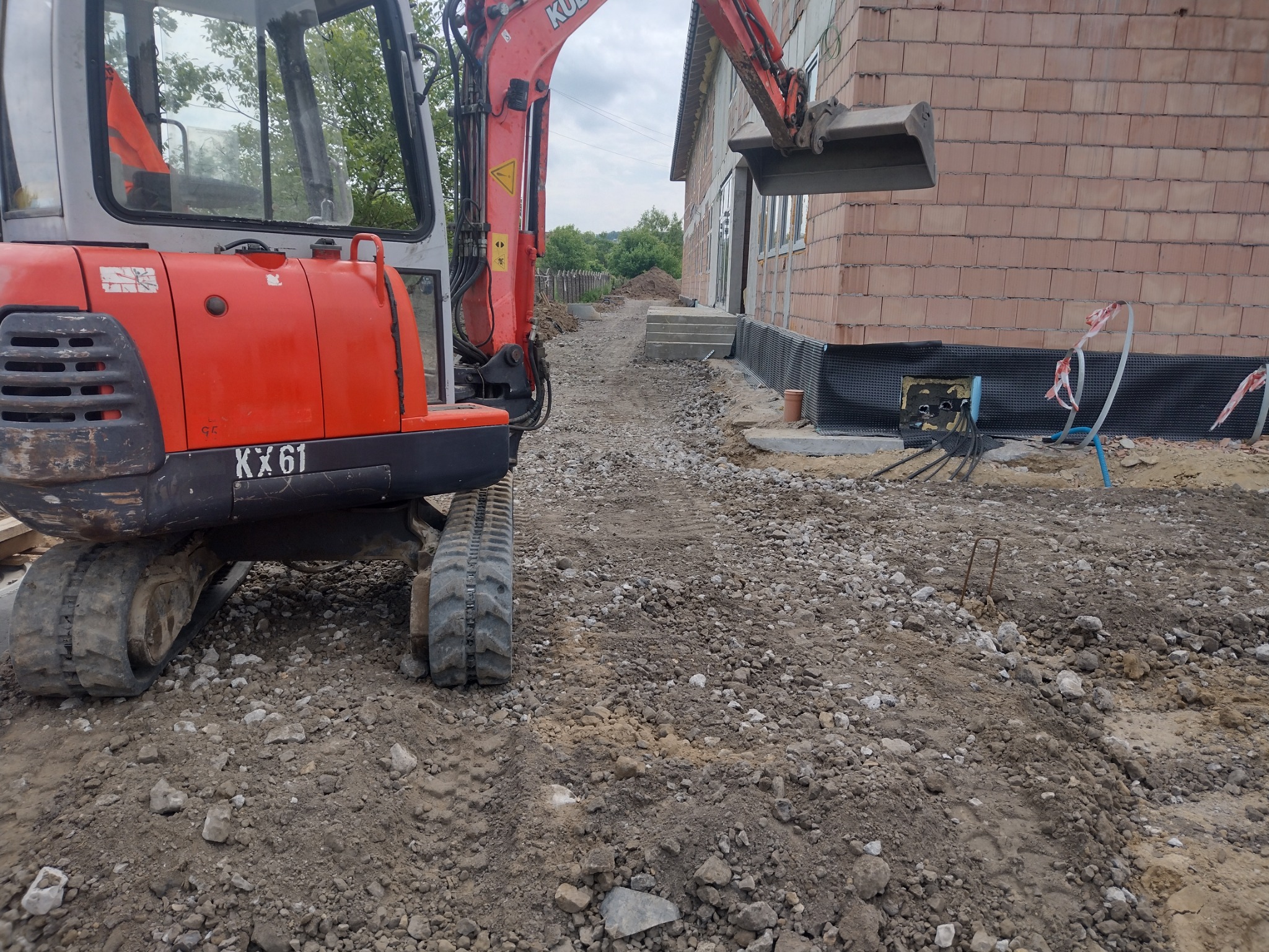 Czerwona mini koparka Kubota KX61 na placu budowy, widoczny fragment budynku z cegły, przygotowane przyłącza i izolacja fundamentów.
