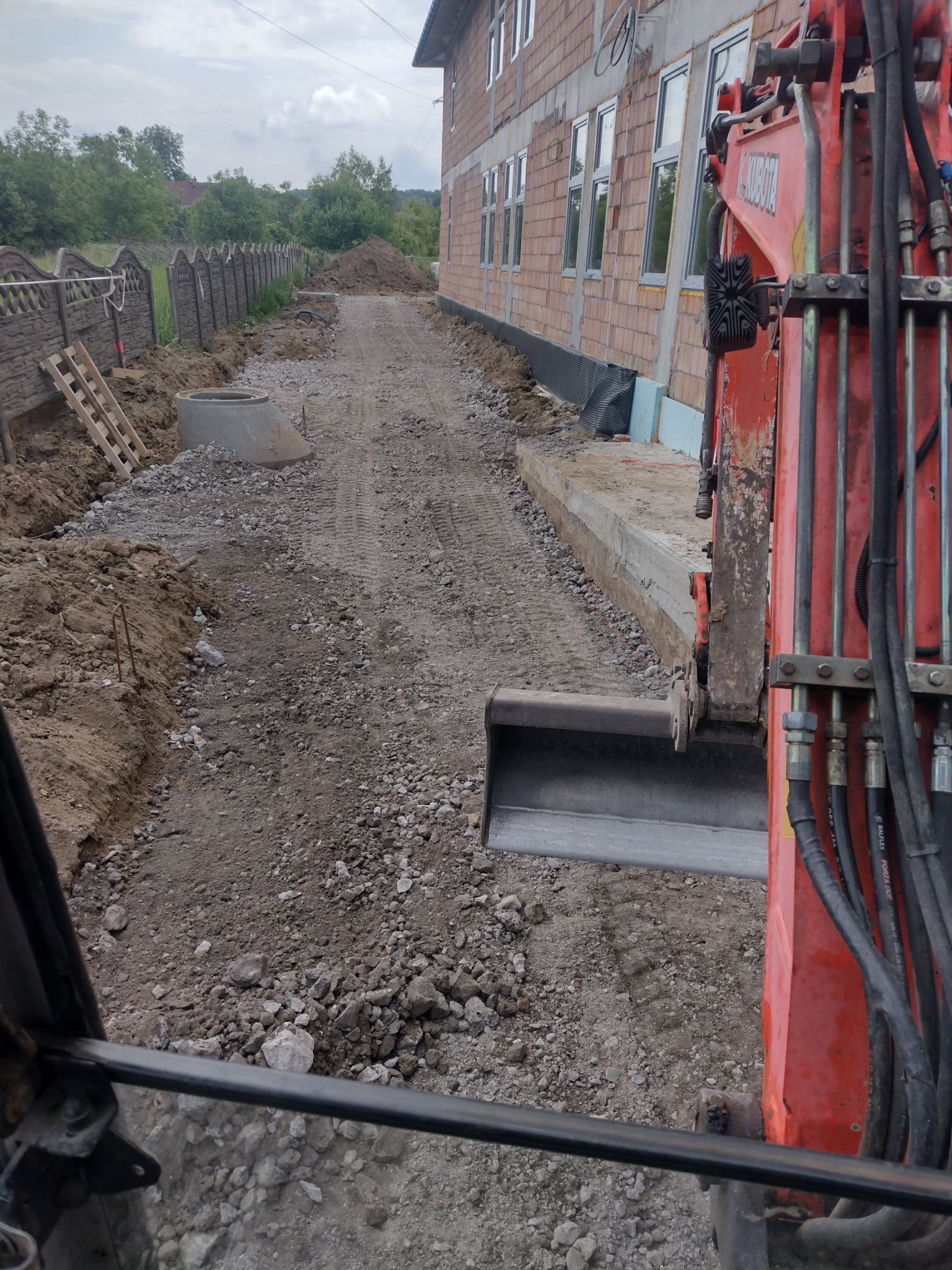 Czerwona koparka Kubota z łyżką, widok z kabiny operatora na teren budowy z fundamentami budynku w trakcie realizacji, ułożonymi prefabrykatami betonowymi, ziemią, gruzem i ogrodzeniem panelowym...