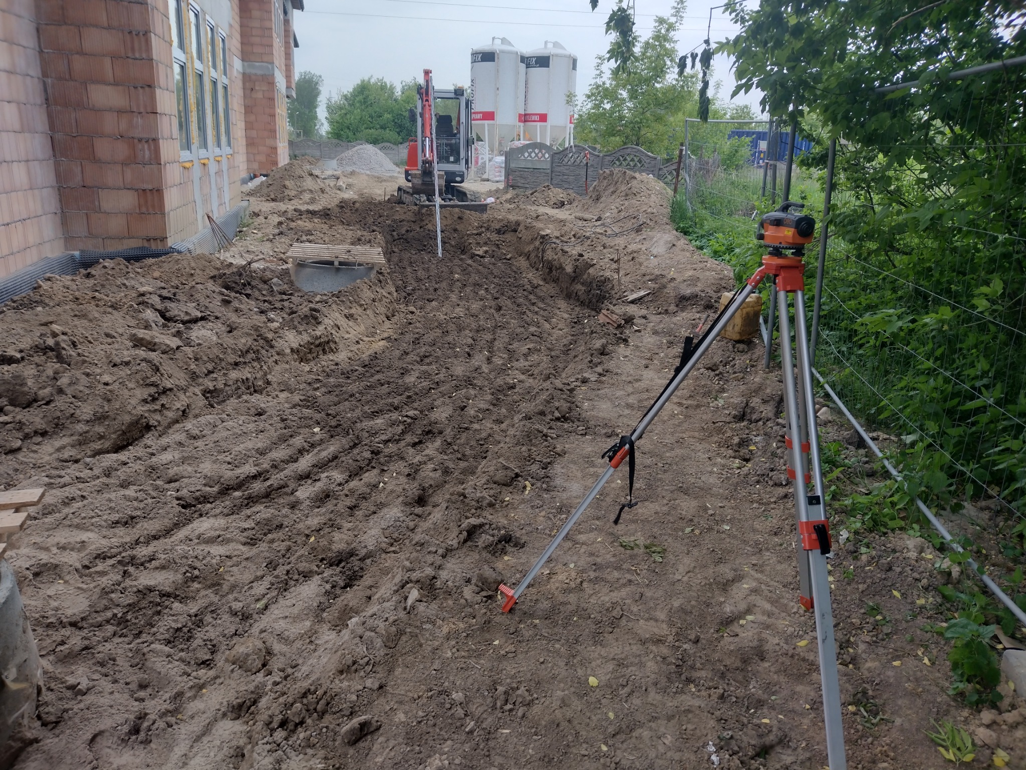 Wykopany rów wzdłuż fundamentów budynku, z widocznym mini-koparką w tle i niwelatorem na statywie na pierwszym planie, teren częściowo wyrównany.