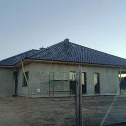 Surowy stan elewacji domu jednorodzinnego z ciemną dachówką, widoczne rusztowania i przygotowania do prac wykończeniowych, żółte rury spustowe, teren wokół ogrodzony siatką.