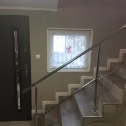 Klatka schodowa z drewnopodobnymi panelami na stopniach i chromowaną balustradą, widok na okno z koronkową firanką i częściowo otwarte drzwi.