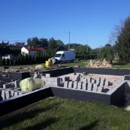 Fundamenty domu w trakcie budowy: wylane ławy fundamentowe, częściowo wymurowane ściany z bloczków betonowych, zabezpieczone czarną izolacją, na placu budowy stoi betoniarka i biały samochód...