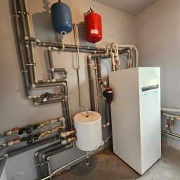 Złożona instalacja grzewcza z białym piecem, zbiornikiem i rurami z izolacją, zamontowana na szarej ścianie w pomieszczeniu technicznym. Widoczne manometry i zawory.