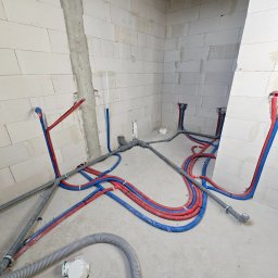 Surowy stan: Instalacja wod-kan w bloku z betonu komórkowego. Czerwone i niebieskie rury prowadzone po podłodze i ścianach, szare rury kanalizacyjne. Widoczne ślady po ołówku.