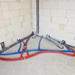 SIDAP ENERGY SPÓŁKA Z OGRANICZONĄ ODPOWIEDZIALNOŚCIĄ - Instalacja ogrzewania podłogowego w surowym wnętrzu: czerwone i niebieskie rury ułożone na szarym betonie, widoczne białe ściany z betonu komórkowego i pionowa rura kanalizacyjna.