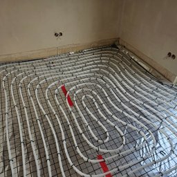 SIDAP ENERGY SPÓŁKA Z OGRANICZONĄ ODPOWIEDZIALNOŚCIĄ - Białe rury ogrzewania podłogowego ułożone na folii z rastrem, widoczne czerwone elementy mocujące, surowy stan ścian i wystające kable elektryczne.