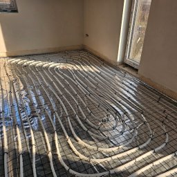 SIDAP ENERGY SPÓŁKA Z OGRANICZONĄ ODPOWIEDZIALNOŚCIĄ - Białe rury ogrzewania podłogowego ułożone na folii i siatce w surowym wnętrzu z oknem. Widoczne mocowania rur do siatki. Promienie słoneczne padają na podłogę.