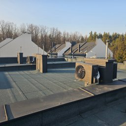 SIDAP ENERGY SPÓŁKA Z OGRANICZONĄ ODPOWIEDZIALNOŚCIĄ - Pompy ciepła na dachu budynku w Warszawie. Widok na kilka jednostek zewnętrznych z szarymi obudowami, zamontowanych na płaskim dachu z widokiem na okoliczne domy.