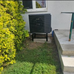 SIDAP ENERGY SPÓŁKA Z OGRANICZONĄ ODPOWIEDZIALNOŚCIĄ - Zamontowana, czarna pompa ciepła Panasonic Aquarea na zewnątrz budynku, obok zielonego krzewu i schodów wejściowych. Widoczny fragment okna i drzwi.