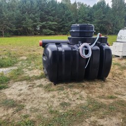 SIDAP ENERGY SPÓŁKA Z OGRANICZONĄ ODPOWIEDZIALNOŚCIĄ - Czarny zbiornik na szambo lub oczyszczalnię ścieków na trawiastym terenie, z białym vanem i paletą bloczków w tle, sugerujący etap instalacji.