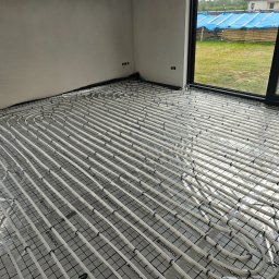 SIDAP ENERGY SPÓŁKA Z OGRANICZONĄ ODPOWIEDZIALNOŚCIĄ - Białe rury ogrzewania podłogowego ułożone na czarnej macie z folią, widoczne otwory na gniazdka w ścianie i okno z widokiem na zewnątrz.