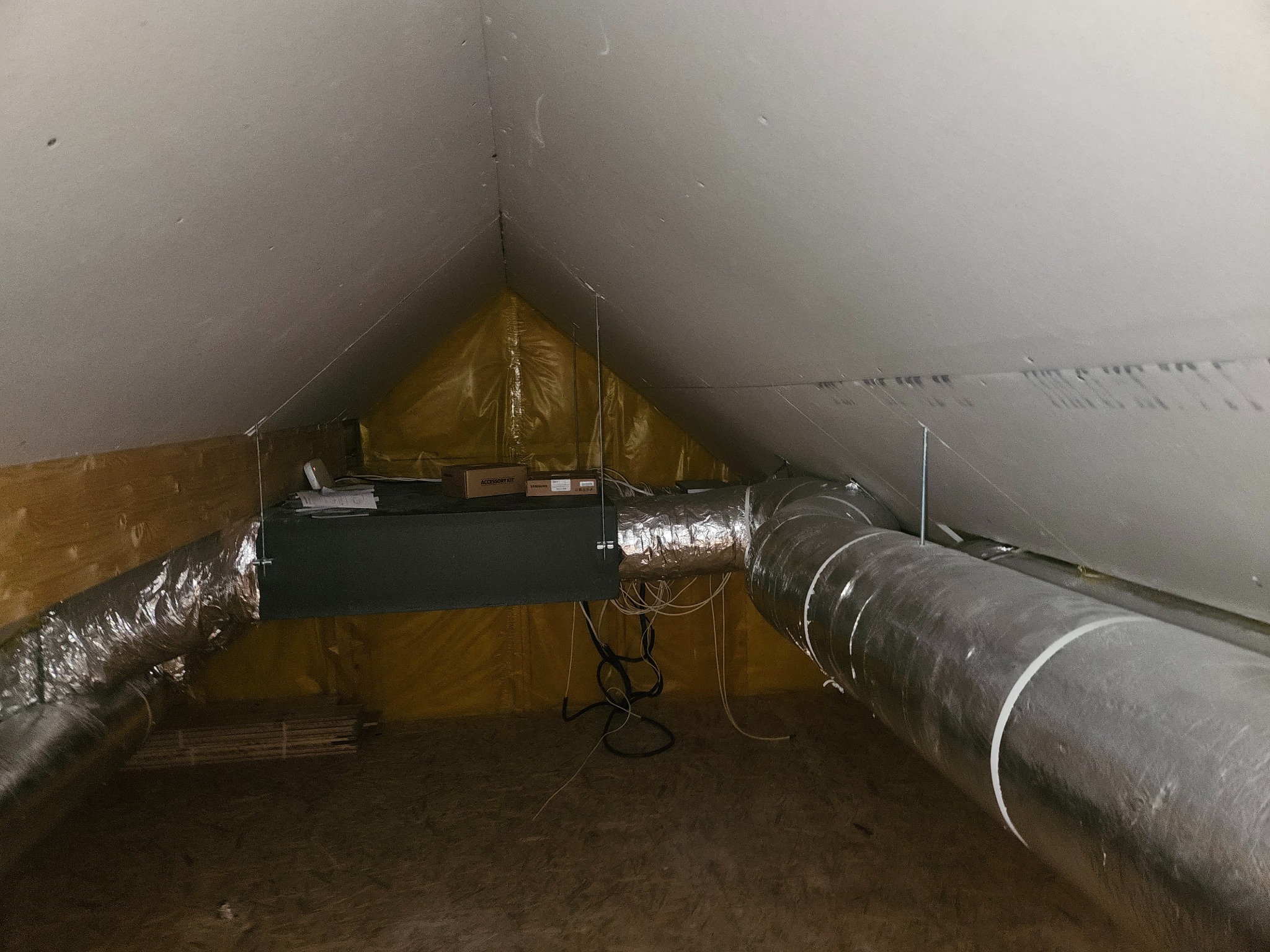 Instalacja wentylacyjna w przestrzeni poddasza: widoczne rury aluminiowe, czarna skrzynka centrali wentylacyjnej z wystającymi kablami oraz elementy konstrukcyjne dachu.