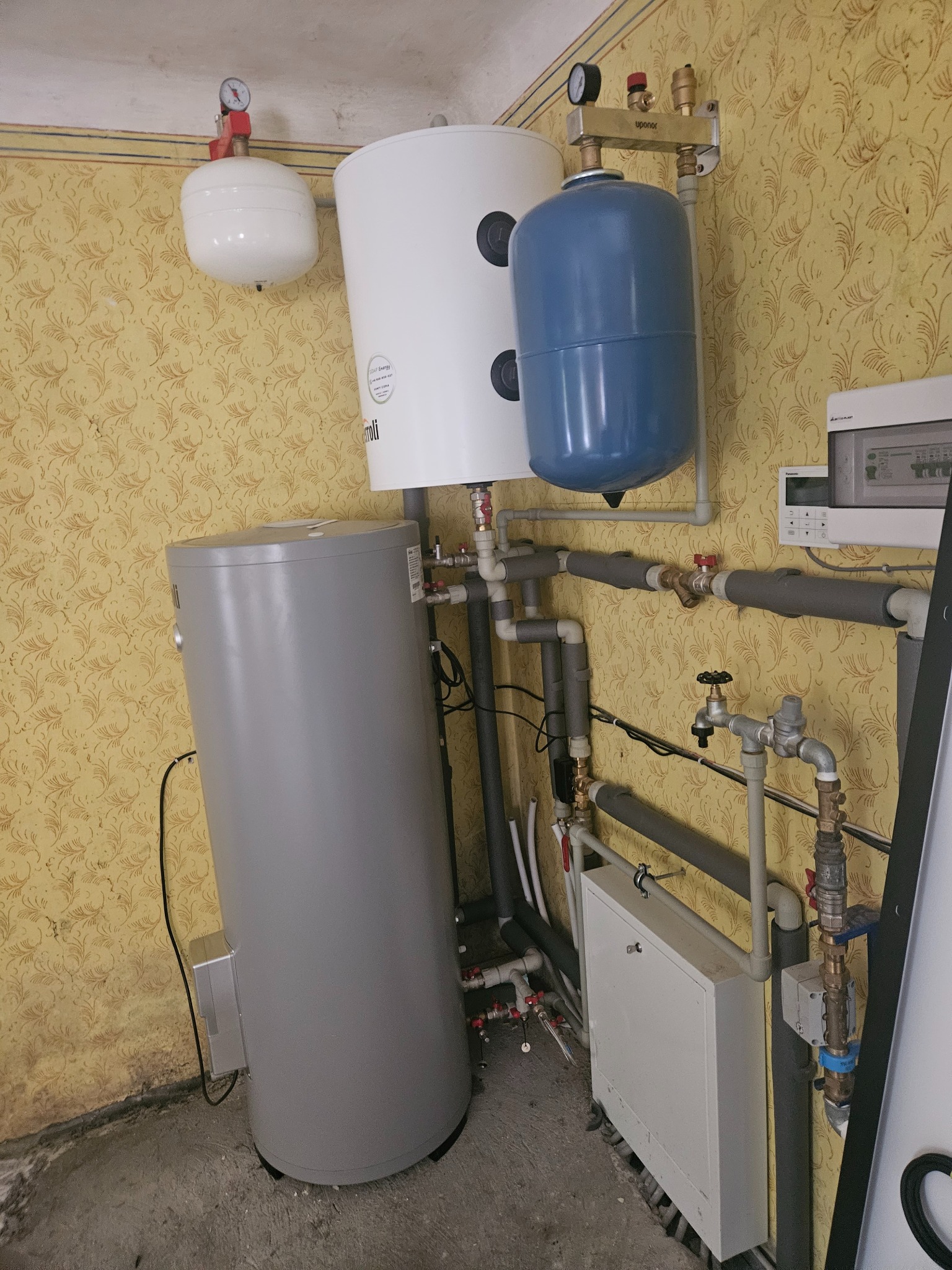 Szary akumulator ciepła, biały zbiornik buforowy i niebieskie naczynie przeponowe zamontowane na ścianie z wzorzystą tapetą, widoczne rury i zawory.