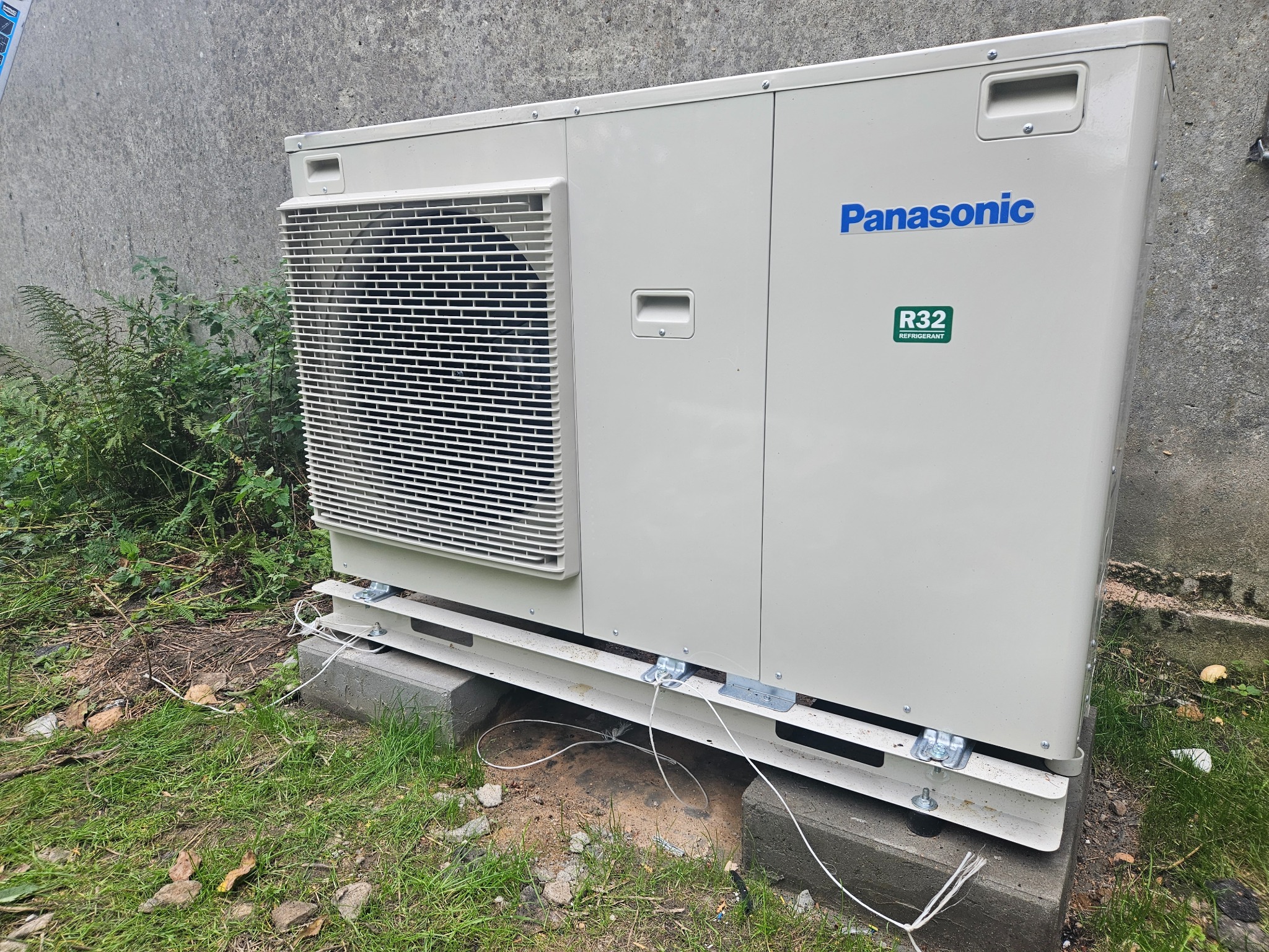 Zewnętrzna jednostka klimatyzacji Panasonic R32 na betonowych podstawach, widoczne przewody instalacyjne, tło stanowi betonowa ściana i fragment roślinności.