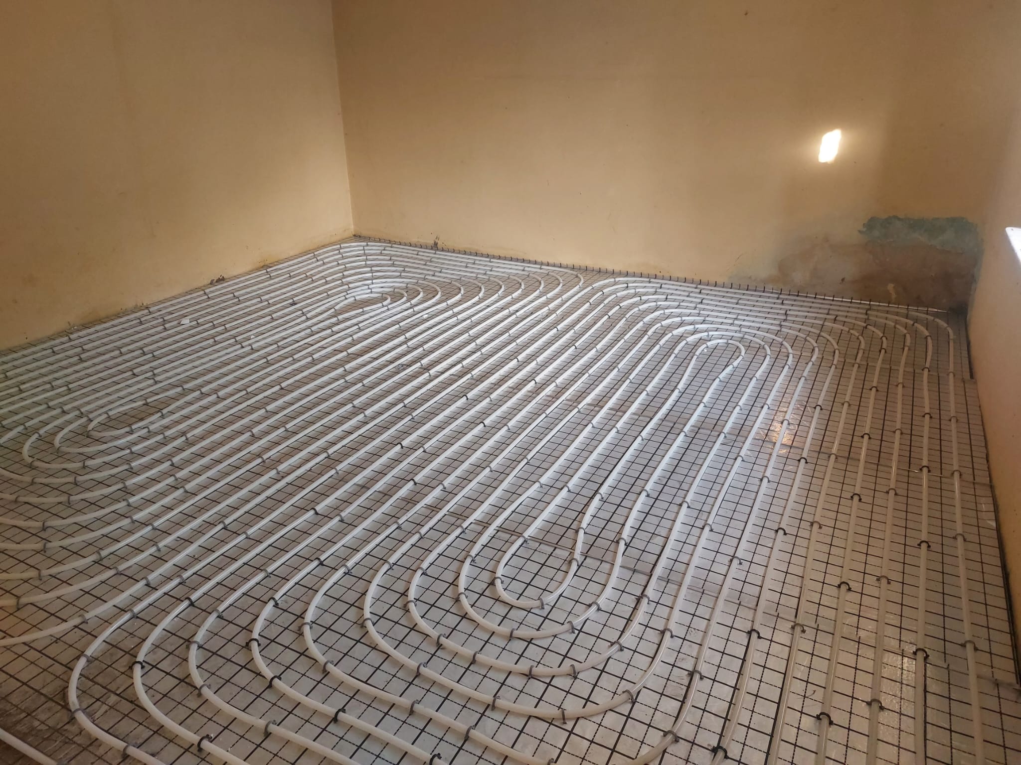 Białe rury ogrzewania podłogowego ułożone w pętle na czarnej siatce w pomieszczeniu o beżowych ścianach, widoczne ślady wilgoci pod sufitem.