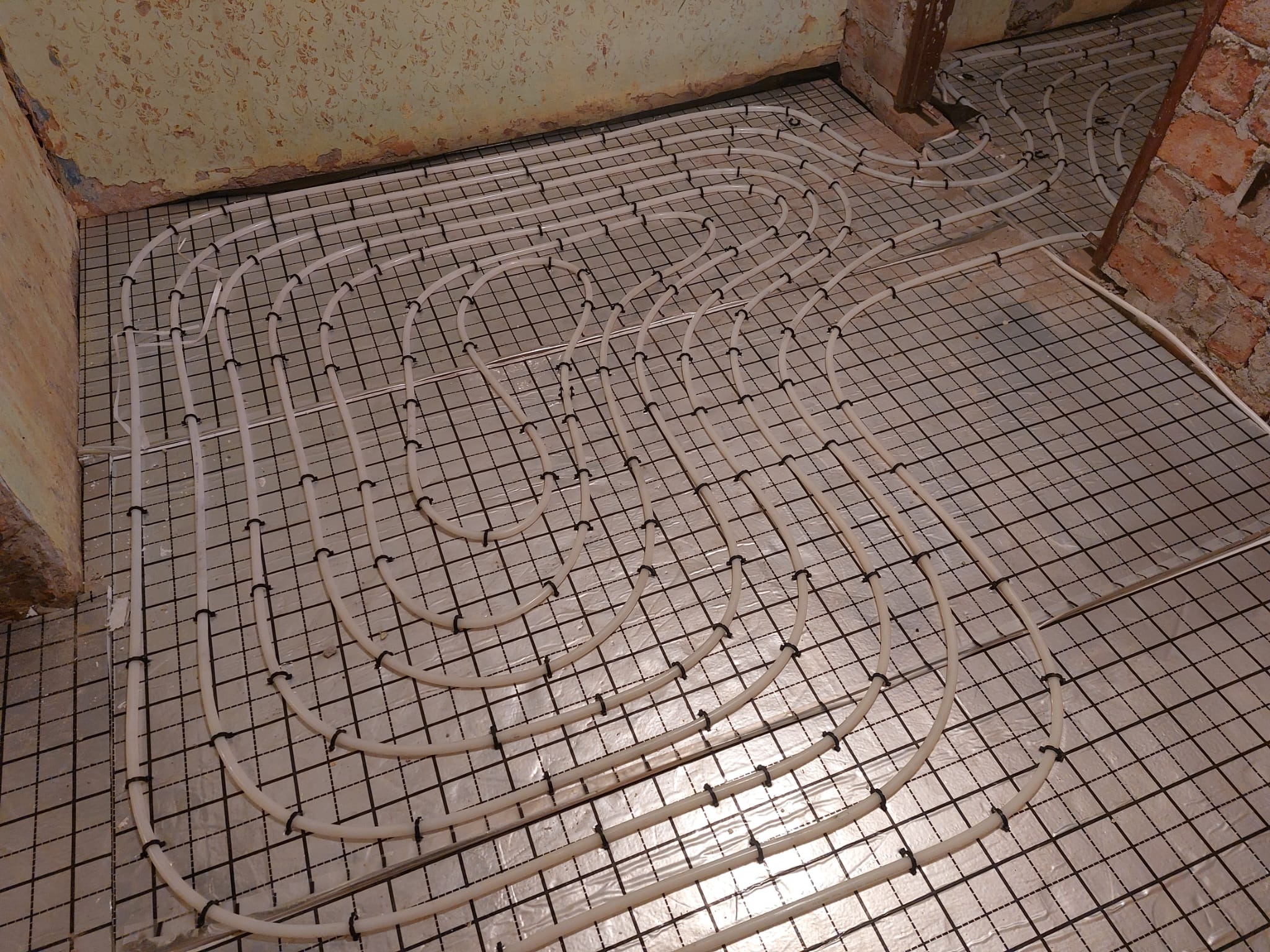 Białe rury ogrzewania podłogowego przymocowane czarnymi klipsami do metalowej siatki na podłożu przed wylewką, widoczne fragmenty ścian z surową cegłą i resztkami farby.