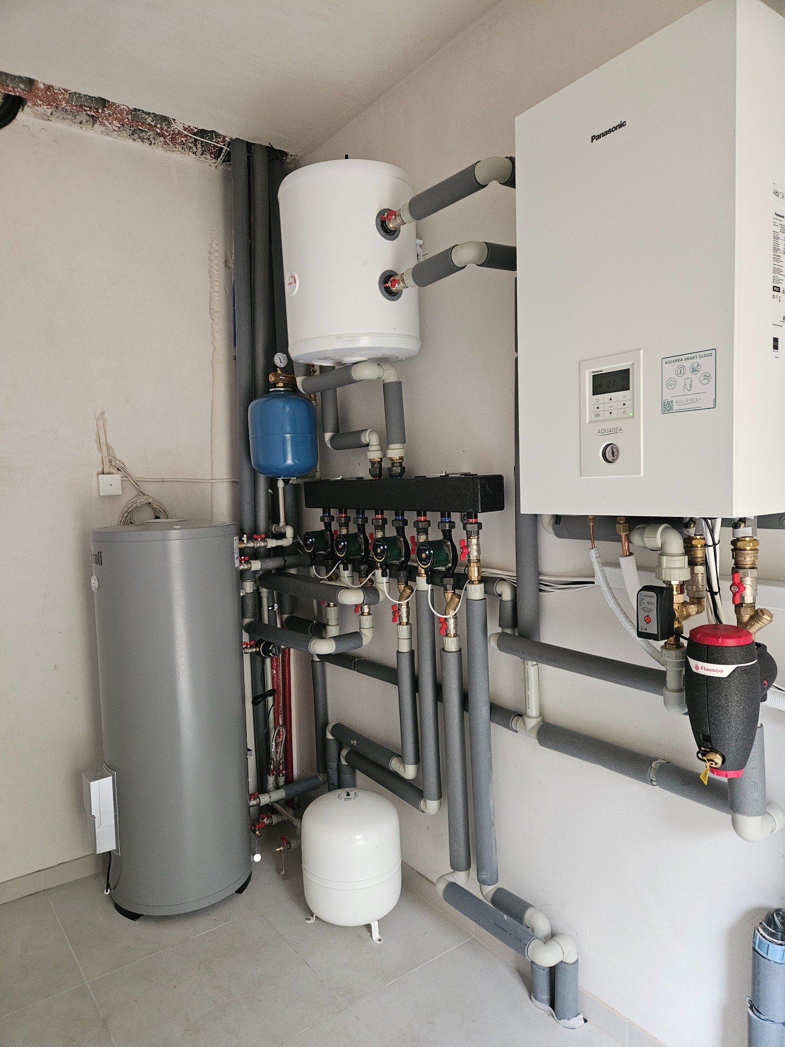 Instalacja grzewcza z zbiornikami buforowymi, pompą obiegową i rozdzielaczem, rury zaizolowane szarą pianką, widoczny manometr i elektroniczny panel sterowania Panasonic Aquarea Smart Cloud.