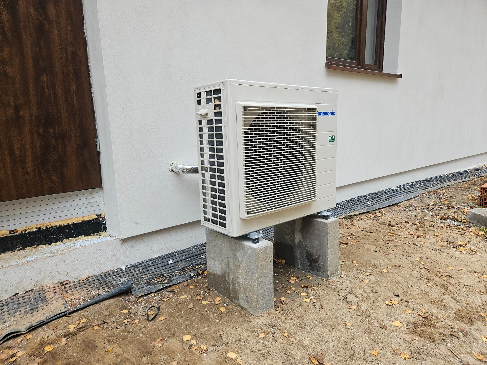 Zewnętrzna jednostka klimatyzacji Panasonic zamontowana na betonowych wspornikach przy ścianie budynku, widoczne rury izolowane i fragment elewacji z oknem oraz liście na ziemi.