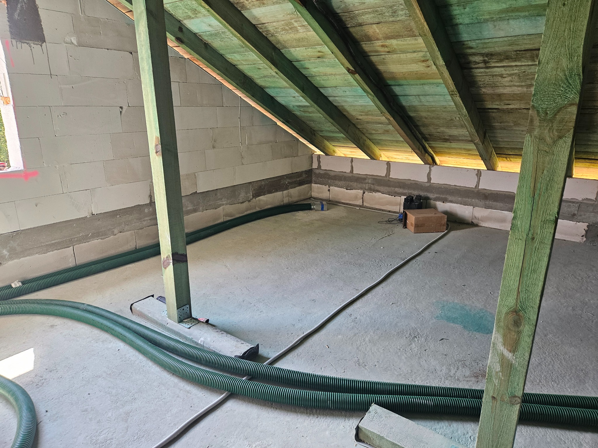 Surowe poddasze z widocznymi elementami konstrukcyjnymi i wstępnie ułożonymi zielonymi rurami wentylacyjnymi, przygotowane do dalszych prac wykończeniowych.