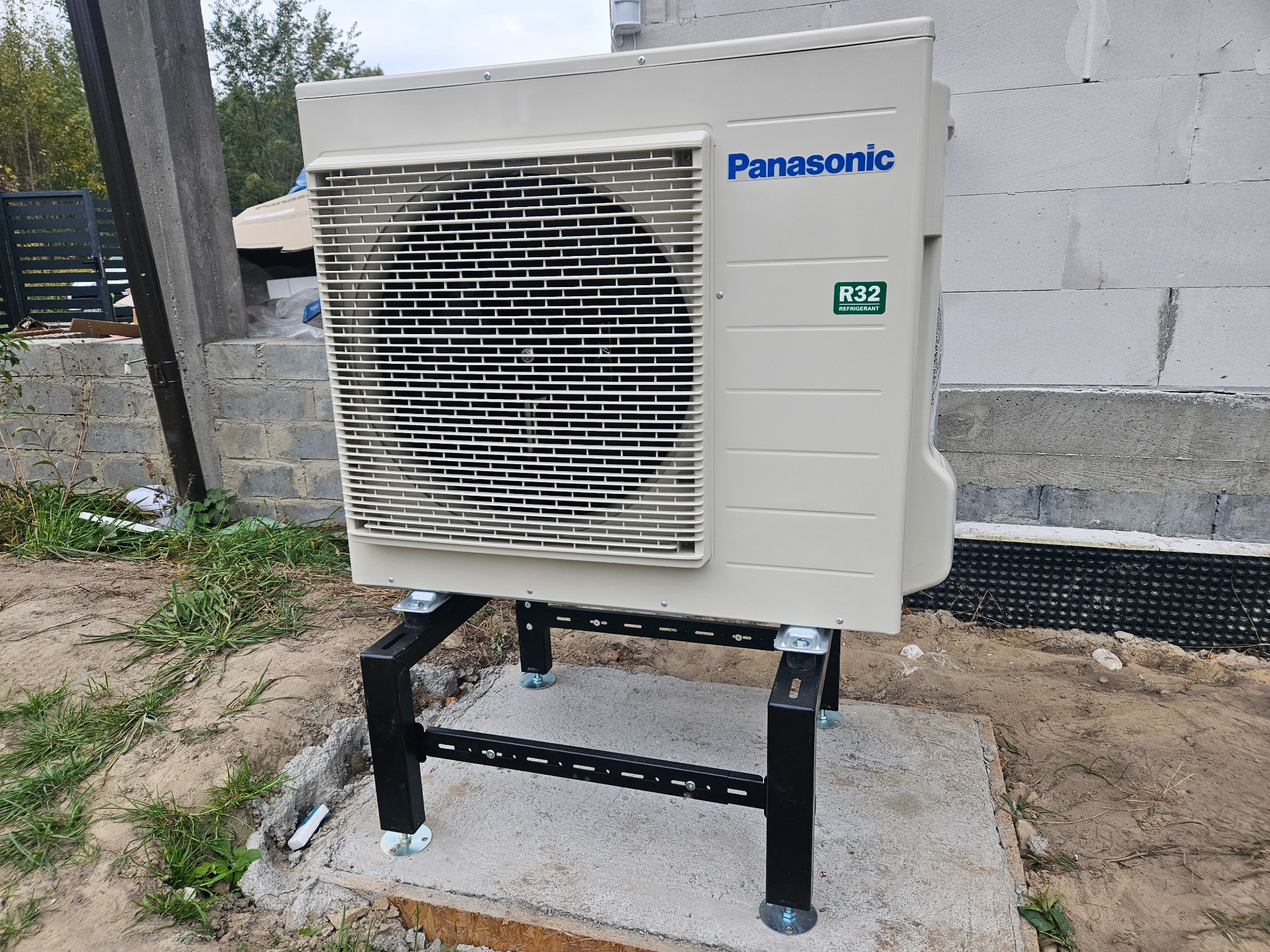 Zewnętrzna jednostka pompy ciepła Panasonic R32 zamontowana na czarnym, stalowym stojaku na betonowej podstawie, przy ścianie budynku z betonu komórkowego.