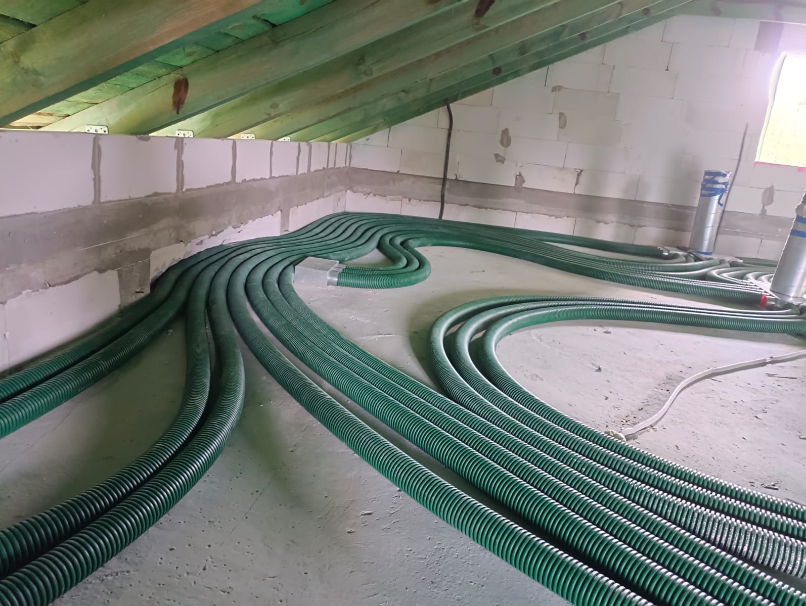 Wnętrze poddasza z ułożonymi zielonymi, karbowanymi rurami wentylacyjnymi, prowadzącymi od ściany z bloczków betonowych w kierunku dalszej części pomieszczenia, widoczne elementy montażowe...