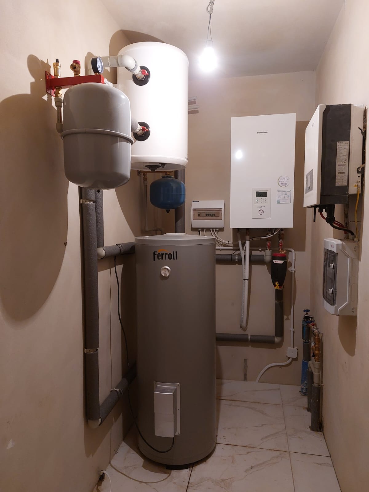 Instalacja grzewcza w pomieszczeniu technicznym z szarym zasobnikiem Ferroli, białym bojlerem, szarą rurą pionową z osprzętem, białym piecem Panasonic i rozdzielnicą elektryczną na ścianie.