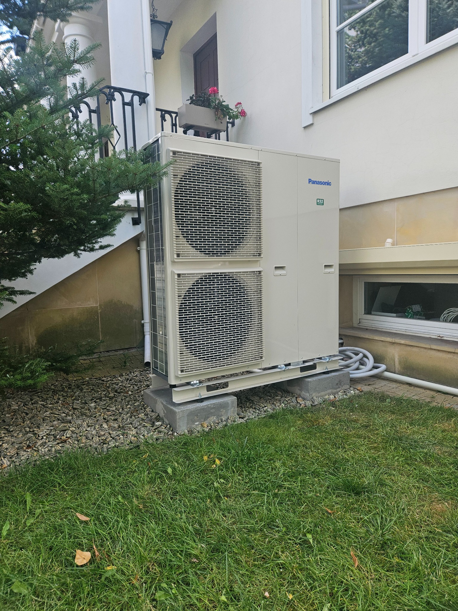 Zewnętrzna jednostka klimatyzacyjna Panasonic, umieszczona na betonowych bloczkach przy budynku z elementami dekoracyjnymi, widoczne przewody i trawnik.