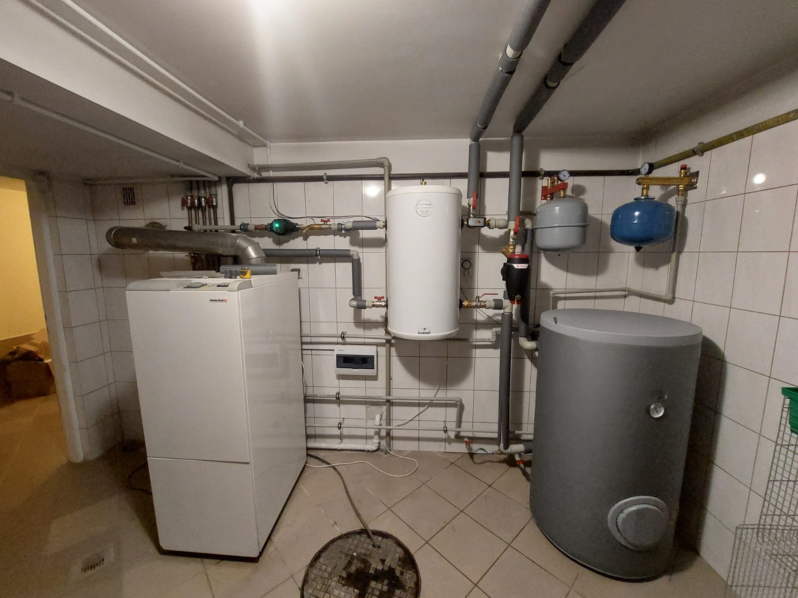Kotłownia z piecem gazowym, zasobnikiem ciepłej wody i naczyniami przeponowymi, widoczna instalacja hydrauliczna z rurami w izolacji oraz kratka ściekowa w podłodze.