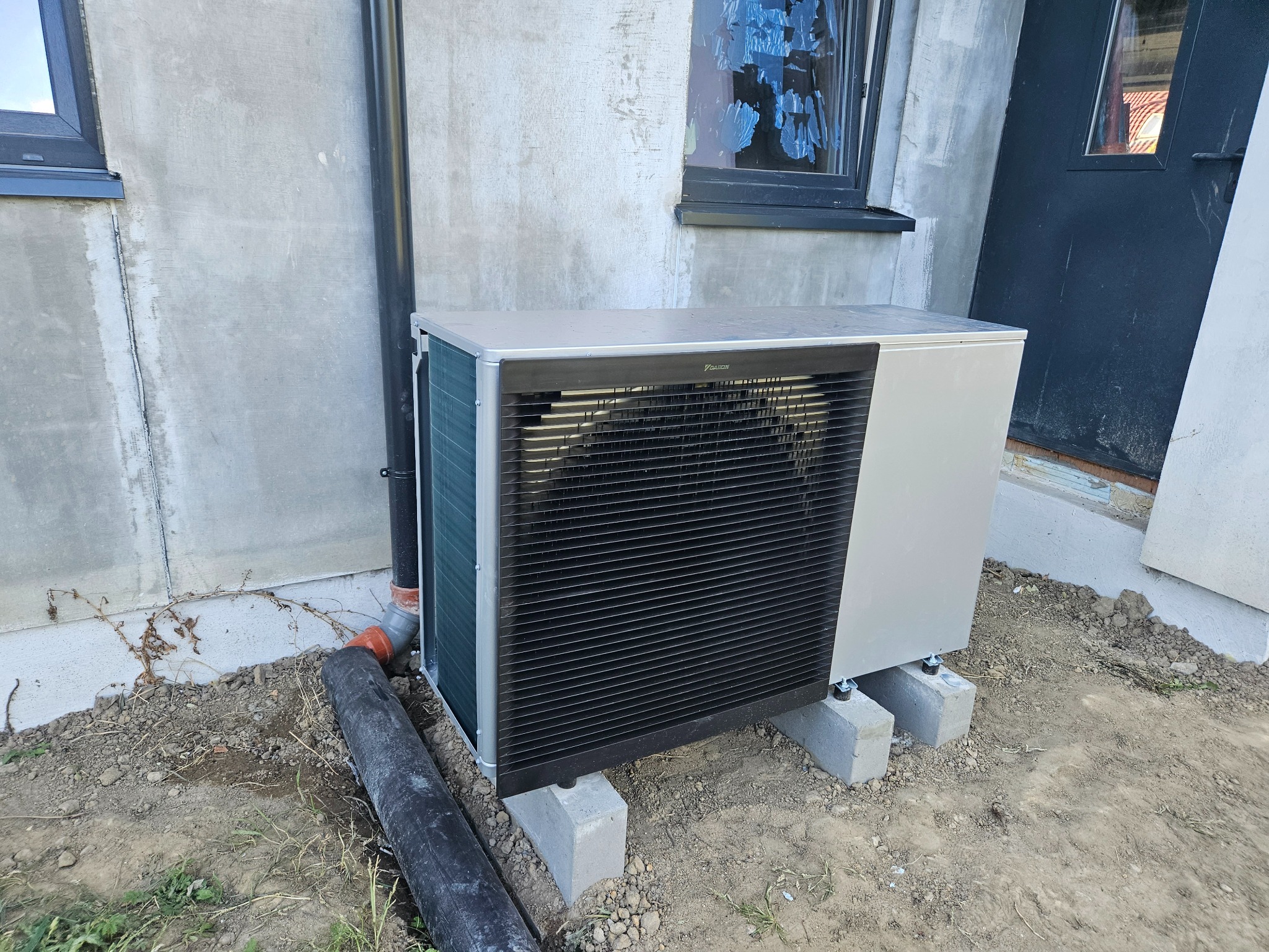 Zewnętrzna jednostka grzewcza Daikin zamontowana na betonowych bloczkach przy szarej ścianie budynku z rurą spustową i oknem z zaklejonymi szybami.