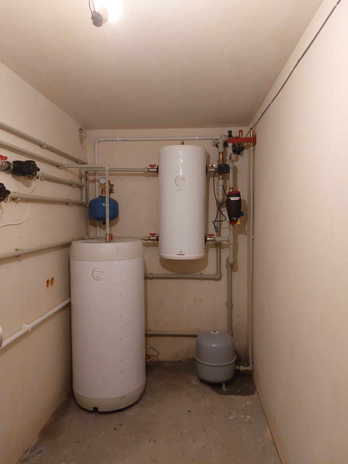 Instalacja z dwoma białymi zbiornikami akumulacyjnymi, niebieskim zbiornikiem wyrównawczym i szarą przeponą w pomieszczeniu z beżowymi ścianami i rurami.