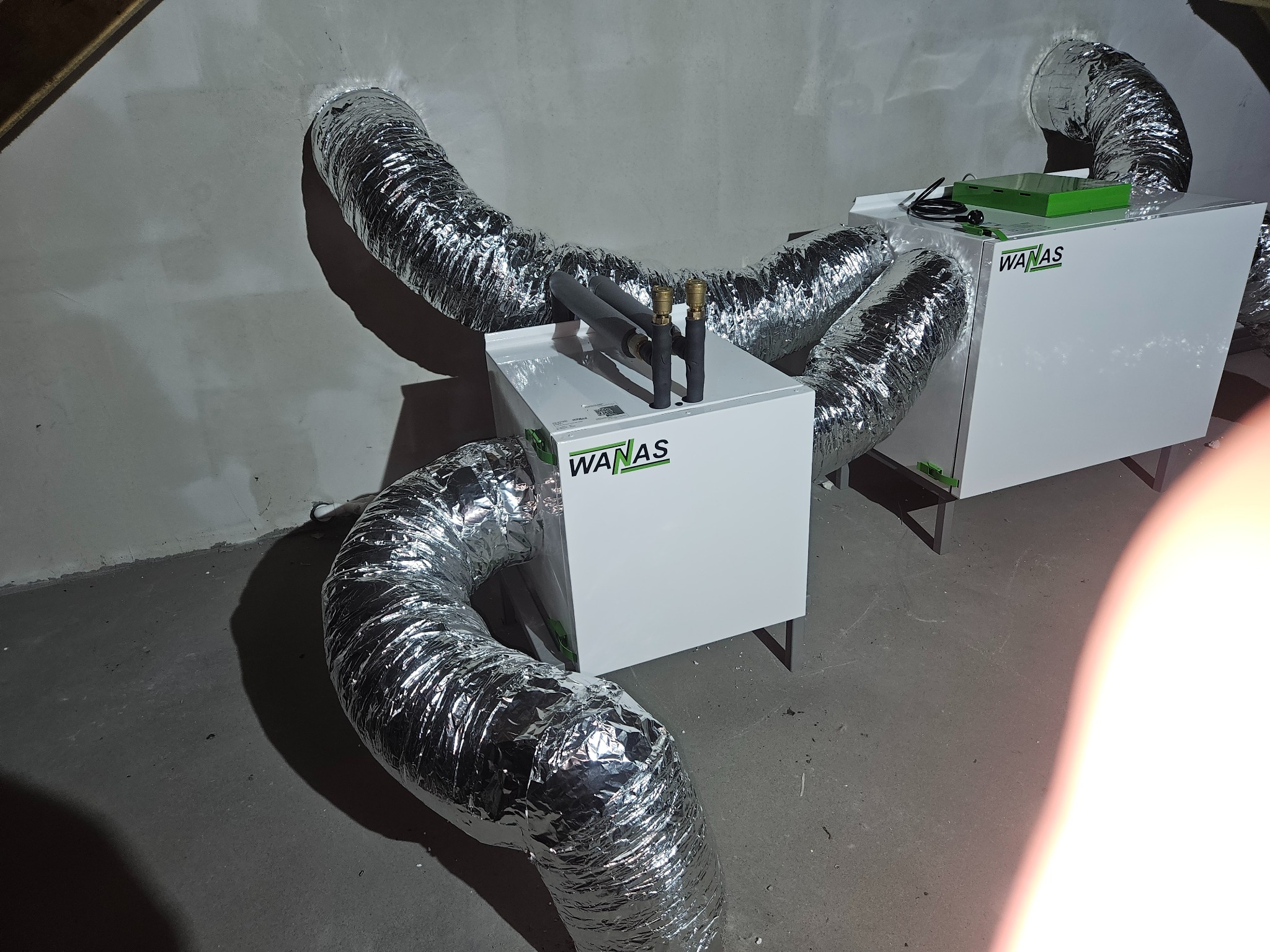 Biała jednostka centralna wentylacyjna z aluminiowymi, elastycznymi przewodami wentylacyjnymi w surowym wnętrzu, widoczne przyłącza hydrauliczne na górze urządzenia, logo WANAS.