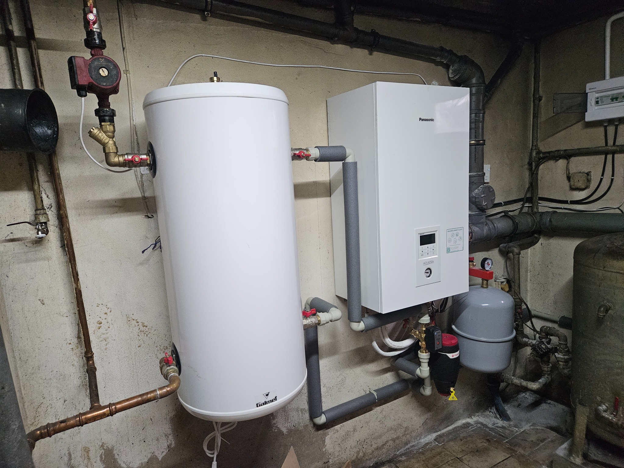 Biały zasobnik ciepłej wody Galmet i wiszący, biały piec Panasonic Aquafea z szarymi rurami w piwnicy. Widoczne miedziane rury, zawory, pompa obiegowa i zbiornik wyrównawczy.