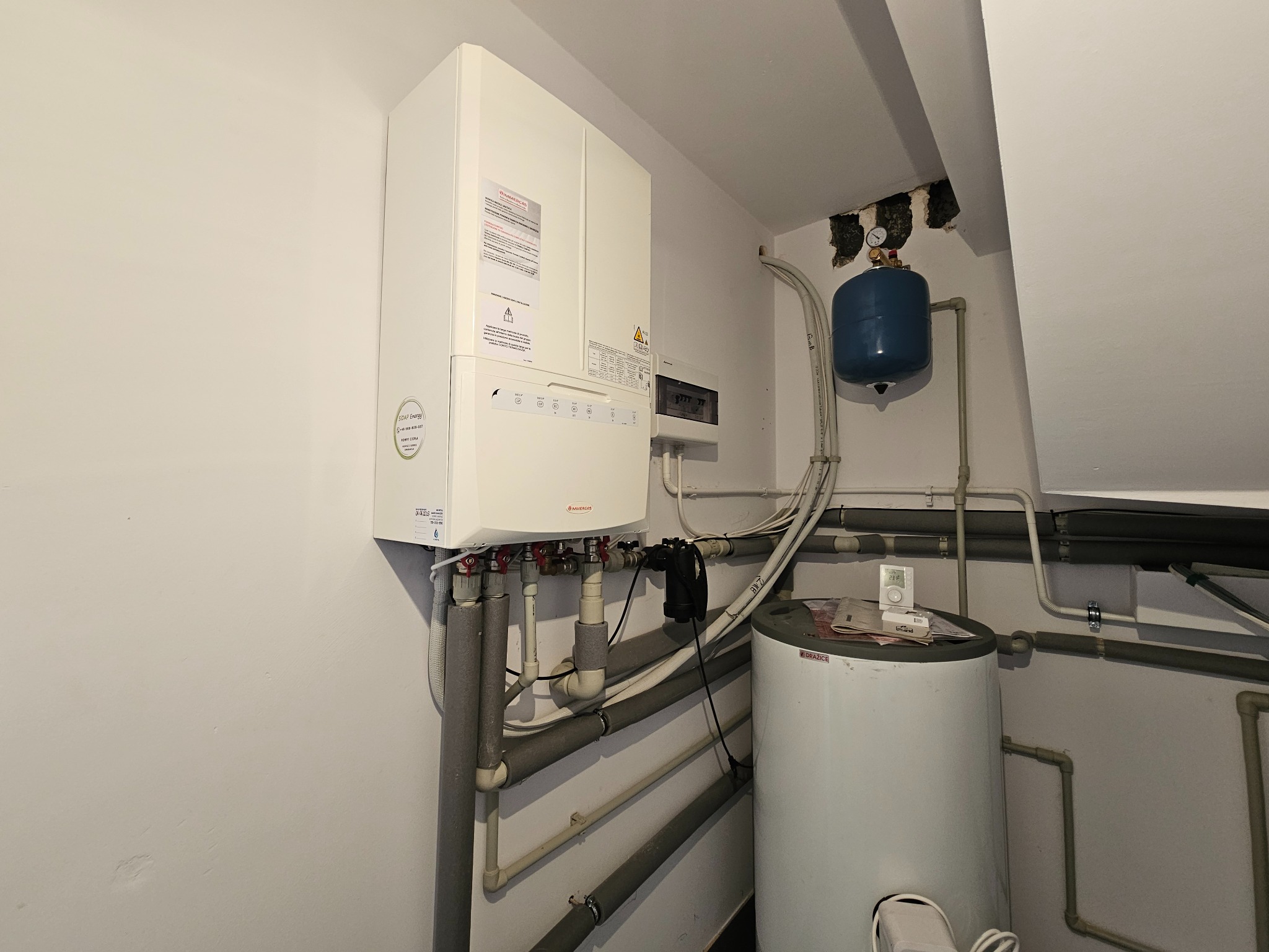 Biały piec gazowy Immergas z podłączonymi rurami, zbiornik wyrównawczy i bojler Drazice w kotłowni z widoczną instalacją hydrauliczną.