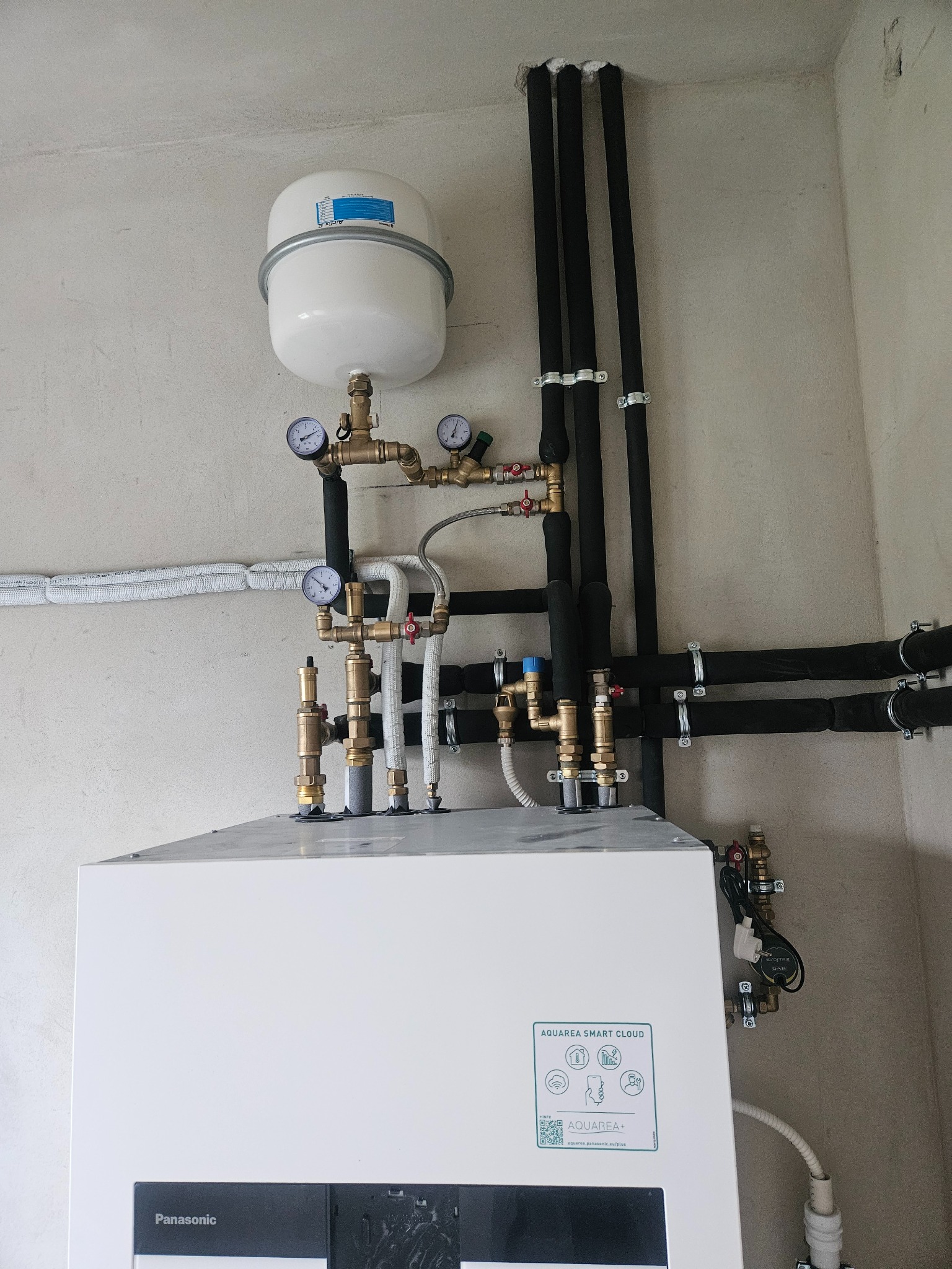 Biała jednostka wewnętrzna z podłączonymi rurami miedzianymi i czarnymi izolowanymi, manometry na górze, zbiornik wyrównawczy, widoczne logo Panasonic Aquarea Smart Cloud