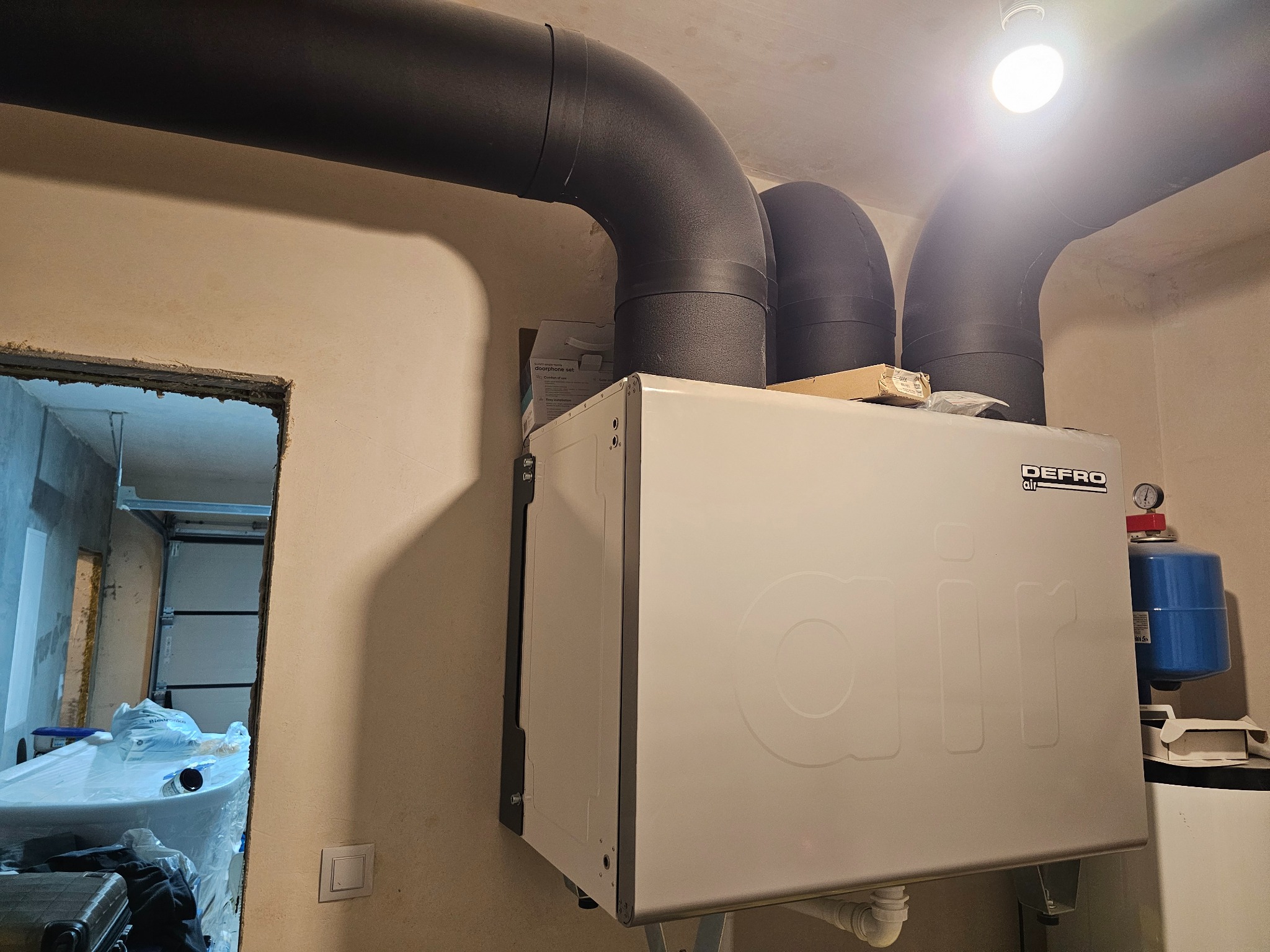 Biała centrala wentylacyjna Defro Air z czarnymi rurami wentylacyjnymi w surowym pomieszczeniu, widoczny manometr i niebieski zbiornik wyrównawczy.