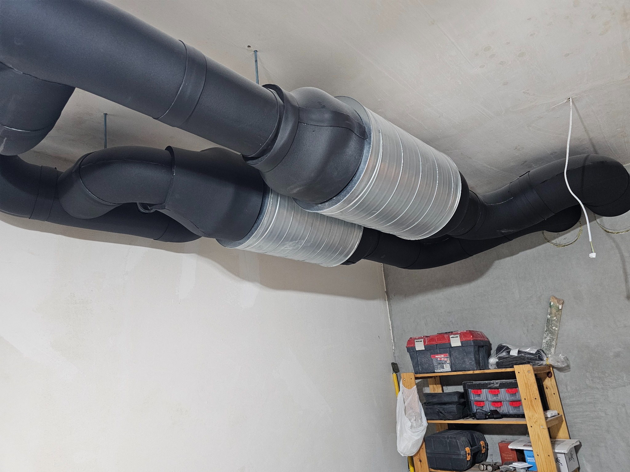 Czarne, zaizolowane rury wentylacyjne z metalowymi elementami łączącymi, zamontowane pod surowym sufitem w pomieszczeniu w trakcie remontu. W tle regał z narzędziami.