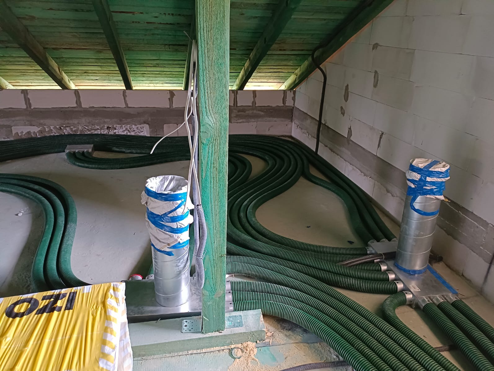 Zielone, karbowane rury wentylacyjne ułożone na podłodze w surowym pomieszczeniu z betonowymi ścianami i drewnianym stropem, widoczne elementy montażowe i izolacyjne.