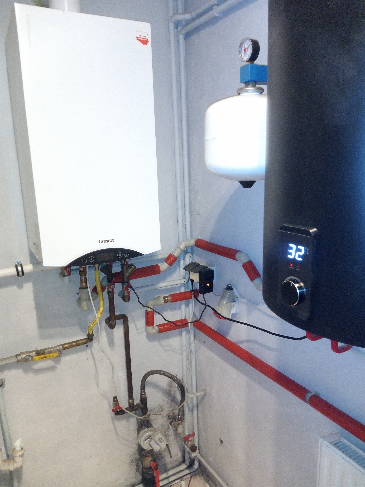 Biały wiszący piec gazowy marki Termet z podłączonymi rurami miedzianymi i plastikowymi w czerwono-białe paski, obok czarny zasobnik ciepłej wody z wyświetlaczem temperatury 32 stopnie Celsjusza...
