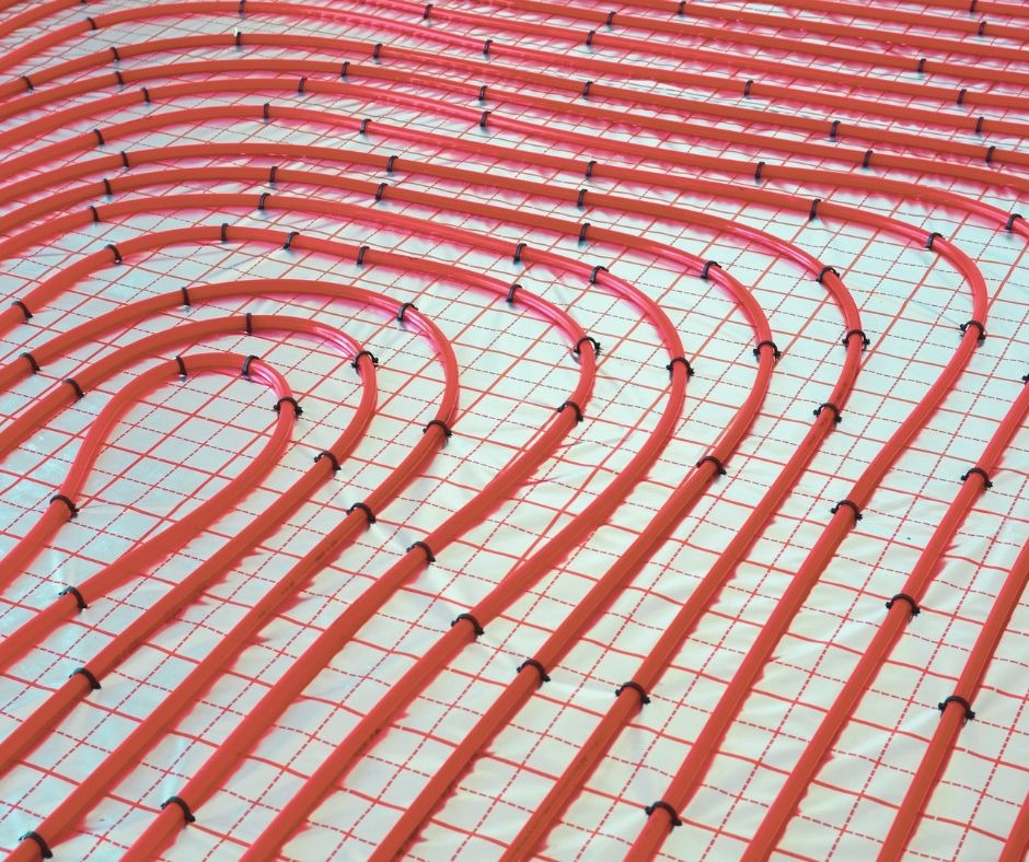 Czerwone rury ogrzewania podłogowego ułożone na białej macie z czerwoną siatką, zamocowane czarnymi klipsami, widok z góry.