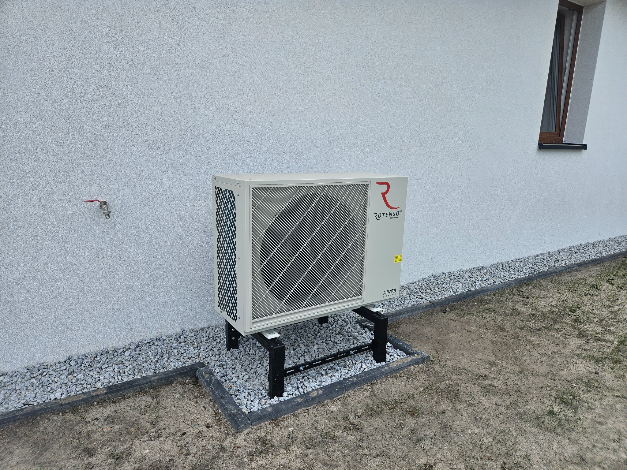 Biała pompa ciepła Rotenso, model Airou, zamontowana na czarnym stelażu przy białej ścianie budynku, otoczona szarym żwirem i obrzeżem.