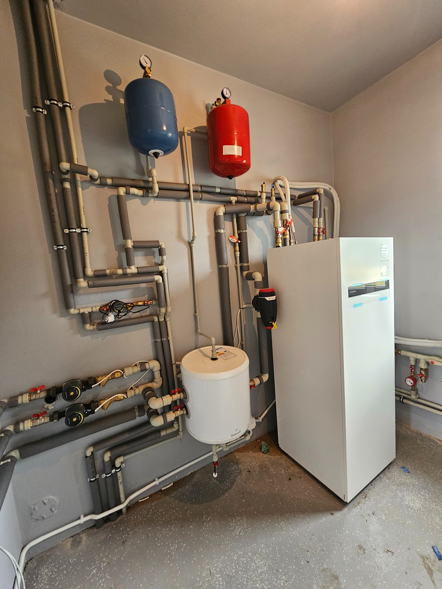 Złożona instalacja grzewcza z białym piecem, zbiornikiem i rurami z izolacją, zamontowana na szarej ścianie w pomieszczeniu technicznym. Widoczne manometry i zawory.