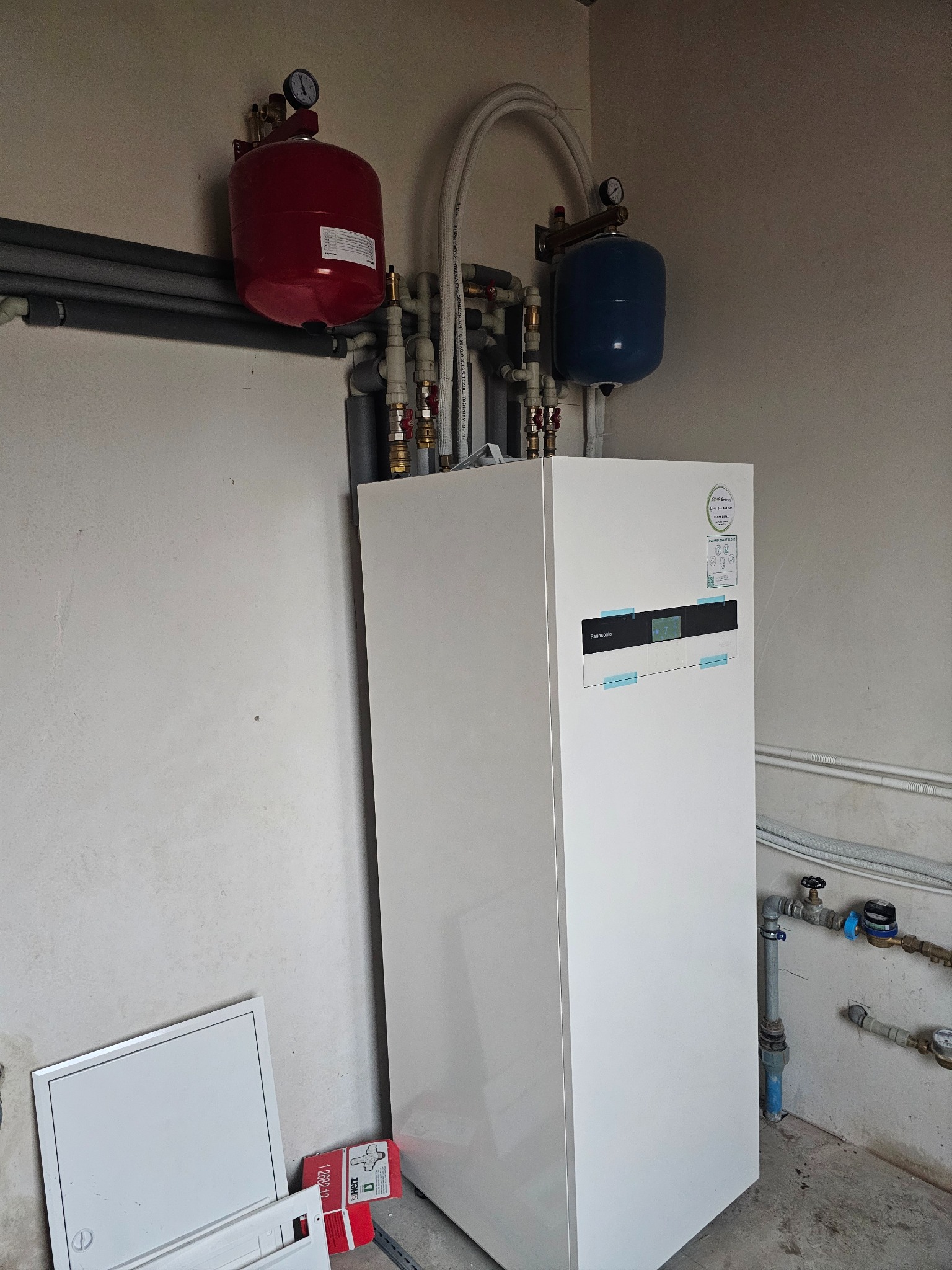 Biała pompa ciepła Panasonic z czerwonym i niebieskim zbiornikiem wyrównawczym, rurami i zaworami na tle szarej ściany. Widoczne elementy instalacji hydraulicznej.