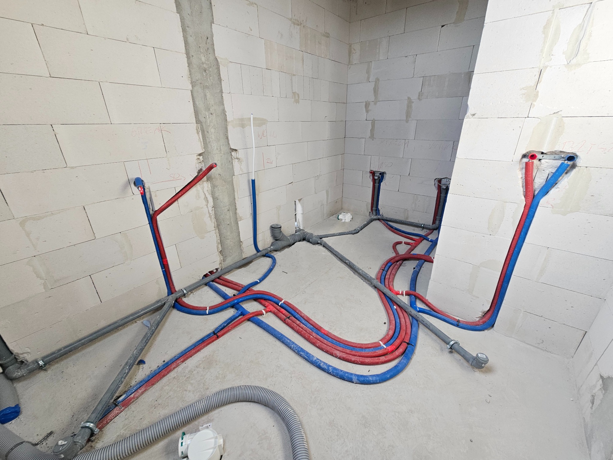 Surowy stan: Instalacja wod-kan w bloku z betonu komórkowego. Czerwone i niebieskie rury prowadzone po podłodze i ścianach, szare rury kanalizacyjne. Widoczne ślady po ołówku.