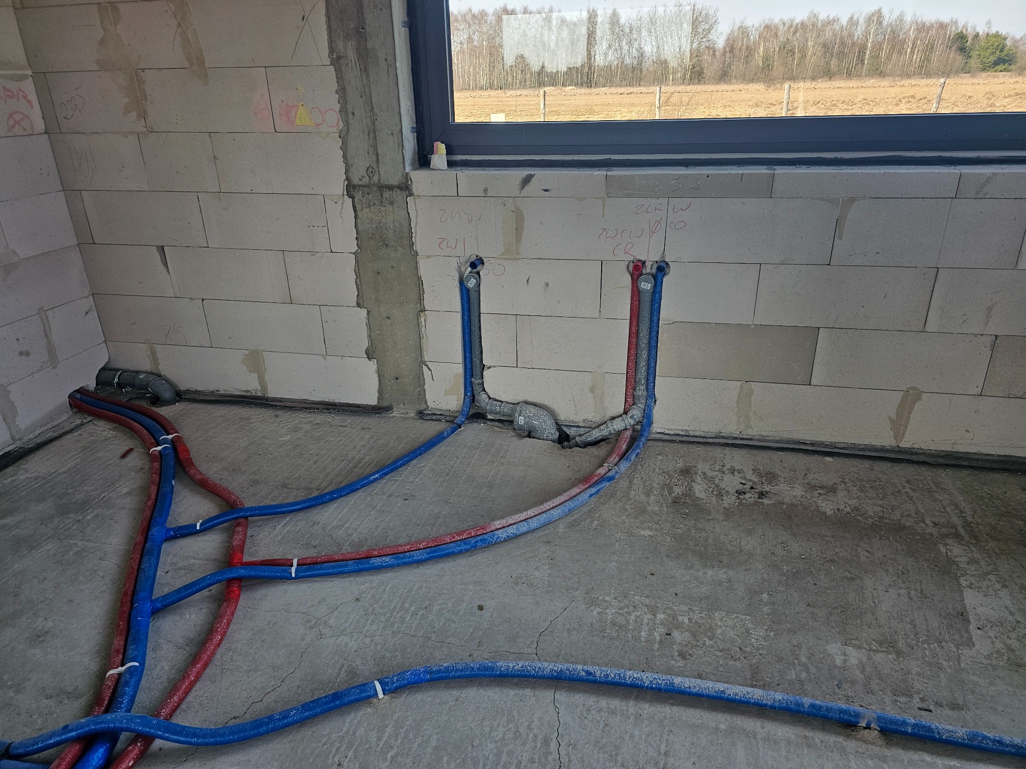 Instalacja wodno-kanalizacyjna w surowym wnętrzu: niebieskie i czerwone rury prowadzone po podłodze, podłączone do wypustów w ścianie z betonu komórkowego pod oknem.