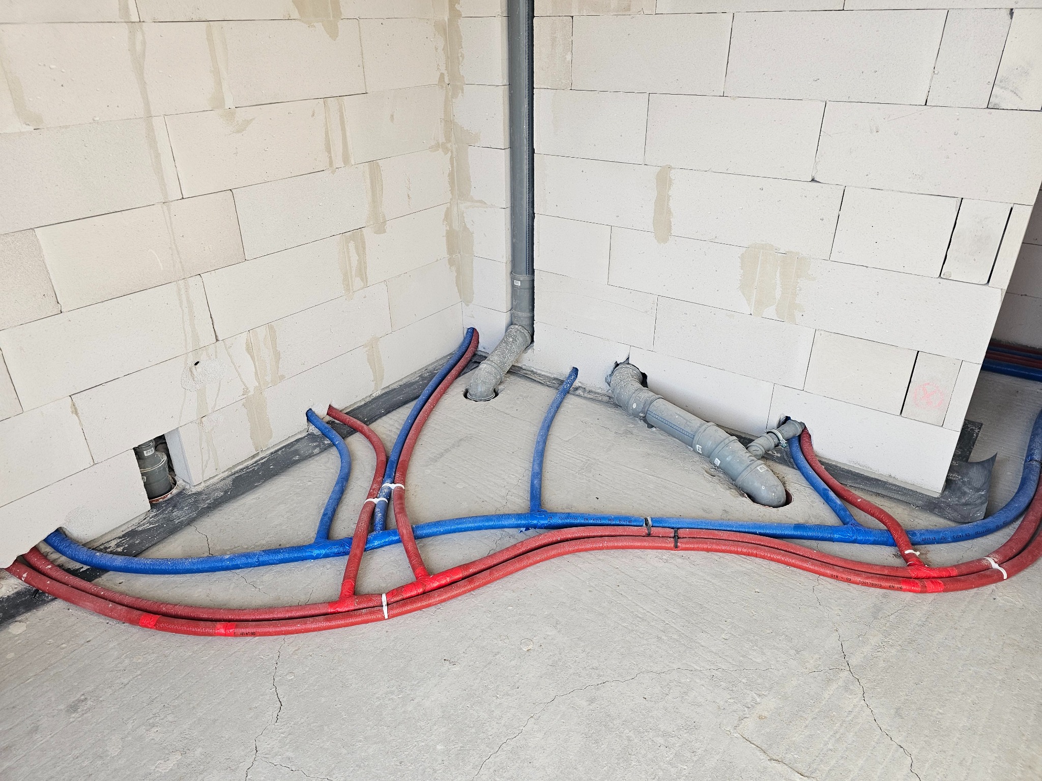 Instalacja ogrzewania podłogowego w surowym wnętrzu: czerwone i niebieskie rury ułożone na szarym betonie, widoczne białe ściany z betonu komórkowego i pionowa rura kanalizacyjna.