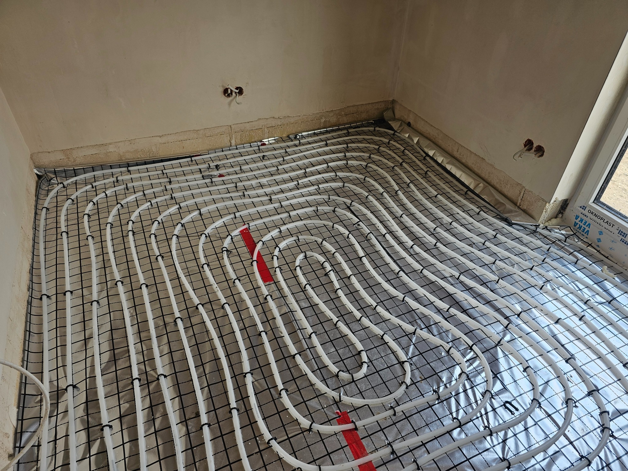 Białe rury ogrzewania podłogowego ułożone na folii z rastrem, widoczne czerwone elementy mocujące, surowy stan ścian i wystające kable elektryczne.