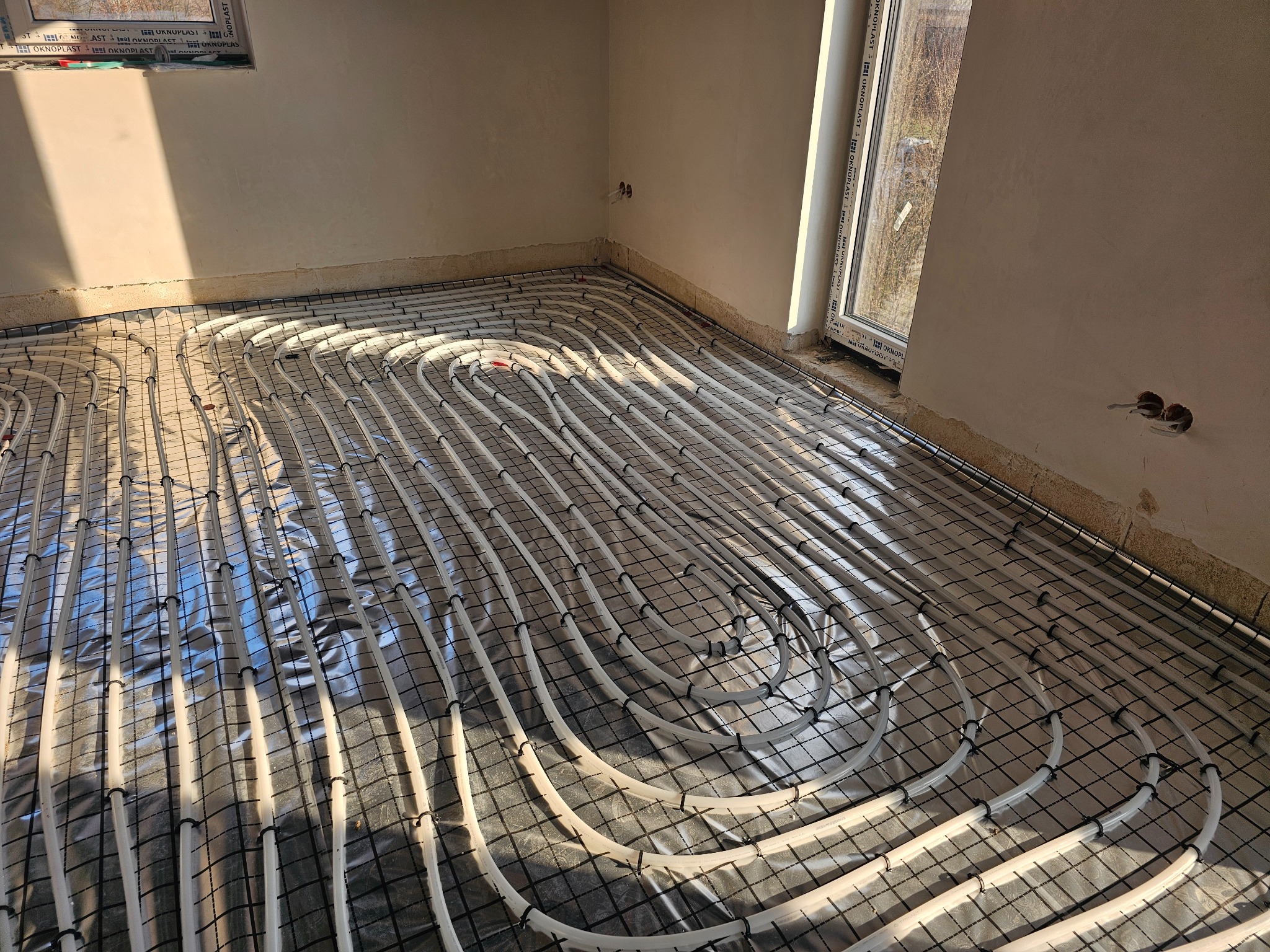 Białe rury ogrzewania podłogowego ułożone na folii i siatce w surowym wnętrzu z oknem. Widoczne mocowania rur do siatki. Promienie słoneczne padają na podłogę.
