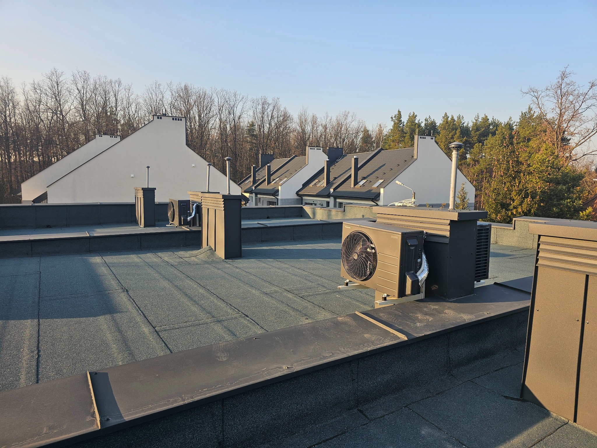 Pompy ciepła na dachu budynku w Warszawie. Widok na kilka jednostek zewnętrznych z szarymi obudowami, zamontowanych na płaskim dachu z widokiem na okoliczne domy.
