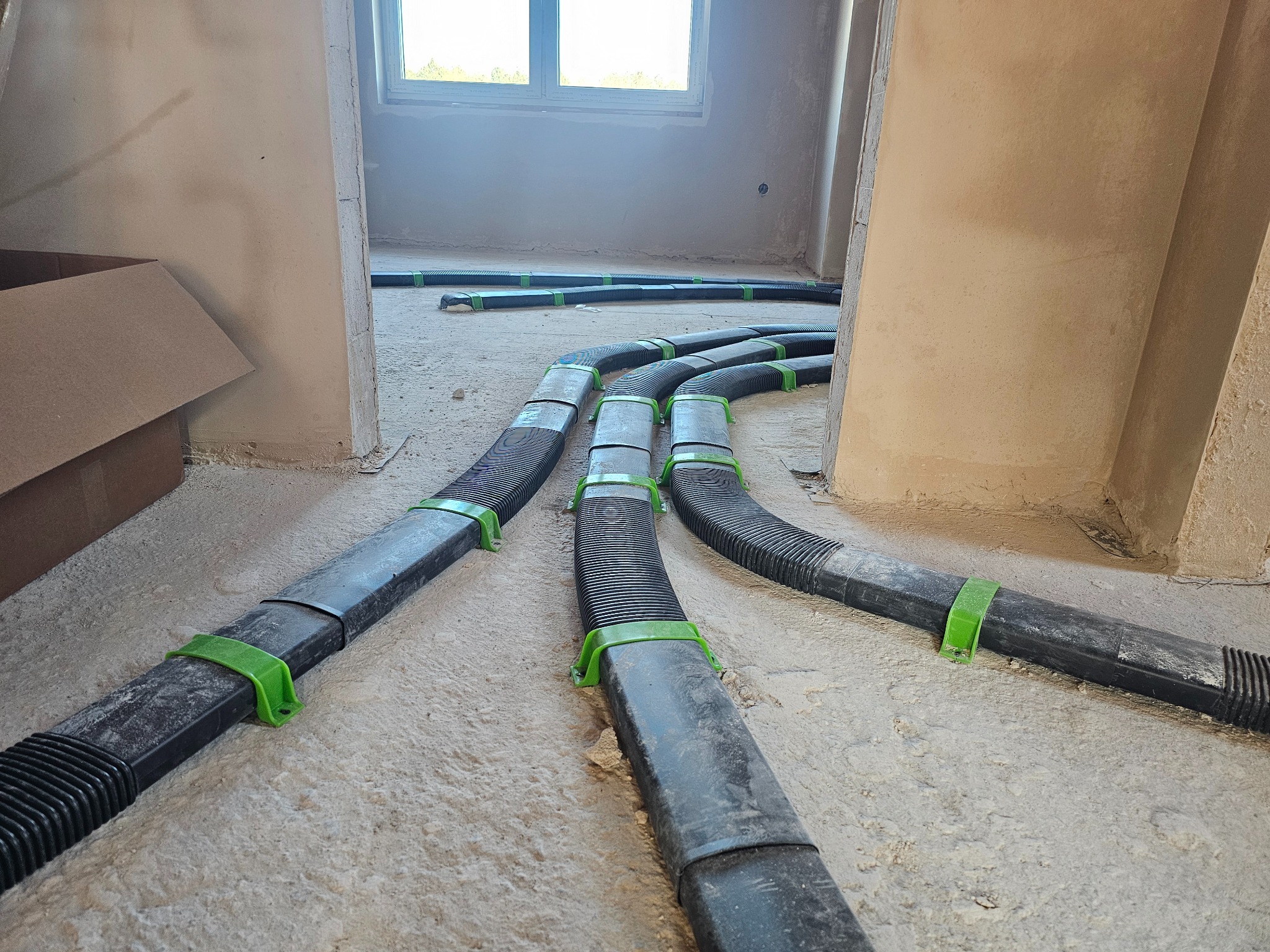Czarny system rur wentylacyjnych z zielonymi uchwytami, ułożony na surowej podłodze w trakcie instalacji w budynku. Widoczne okno w tle i fragment kartonu.