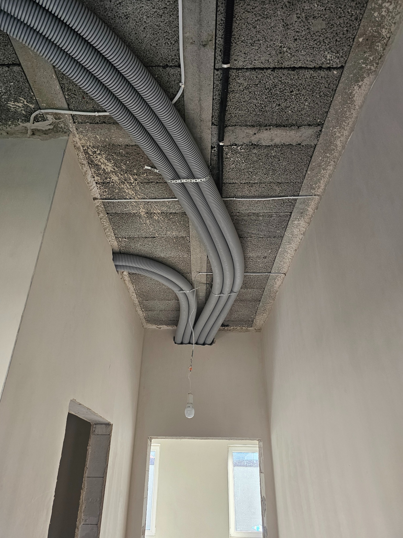 Szare rury wentylacyjne ułożone pod betonowym sufitem w surowym wnętrzu. Widoczne ściany w trakcie wykończenia i surowy otwór drzwiowy.